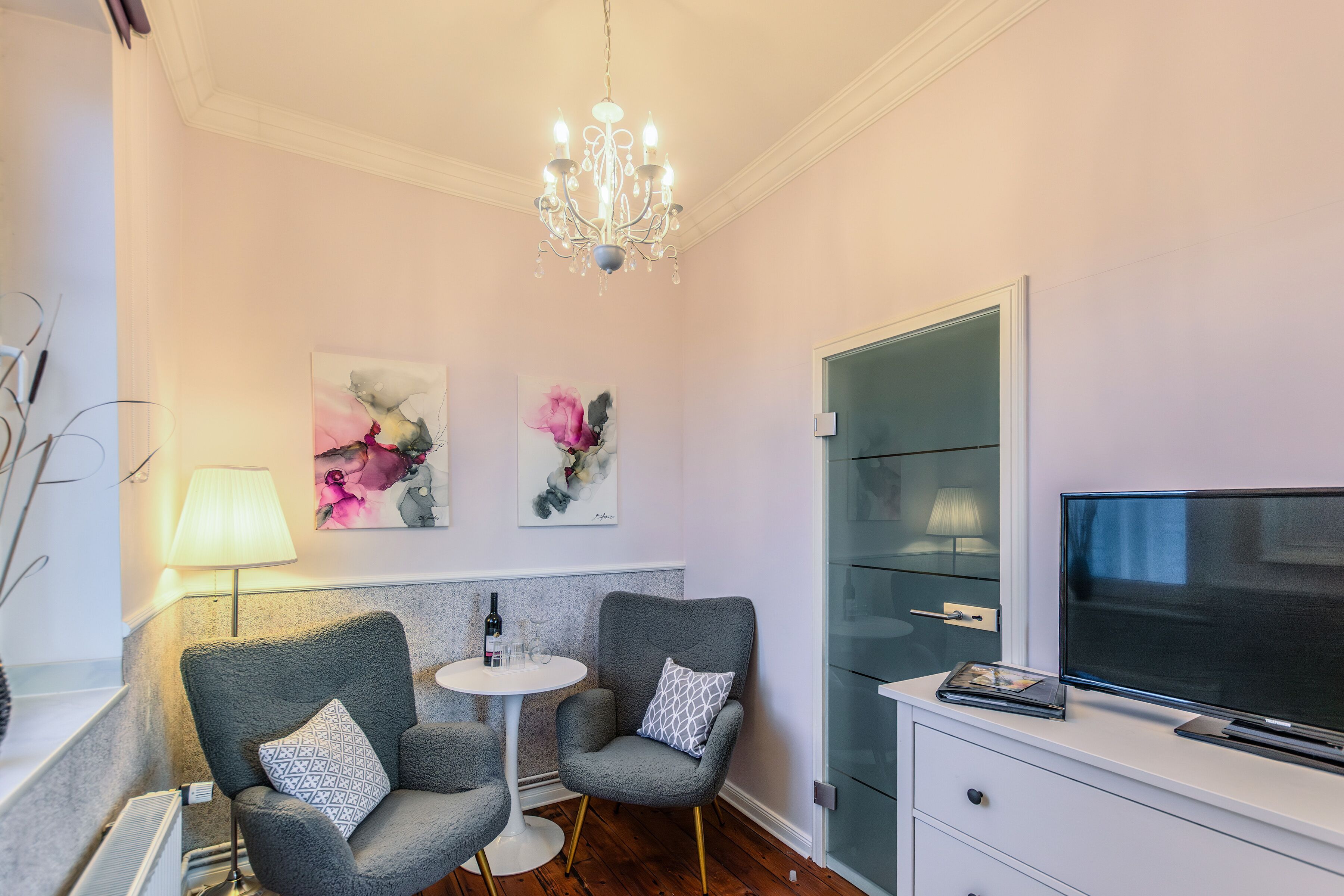 Ein modernes Wohnzimmer mit einer Chandelier, zwei Sesseln, einem kleinen runden Tisch mit einer Flasche, Gläsern und einem Fernseher auf einer Kommode.
