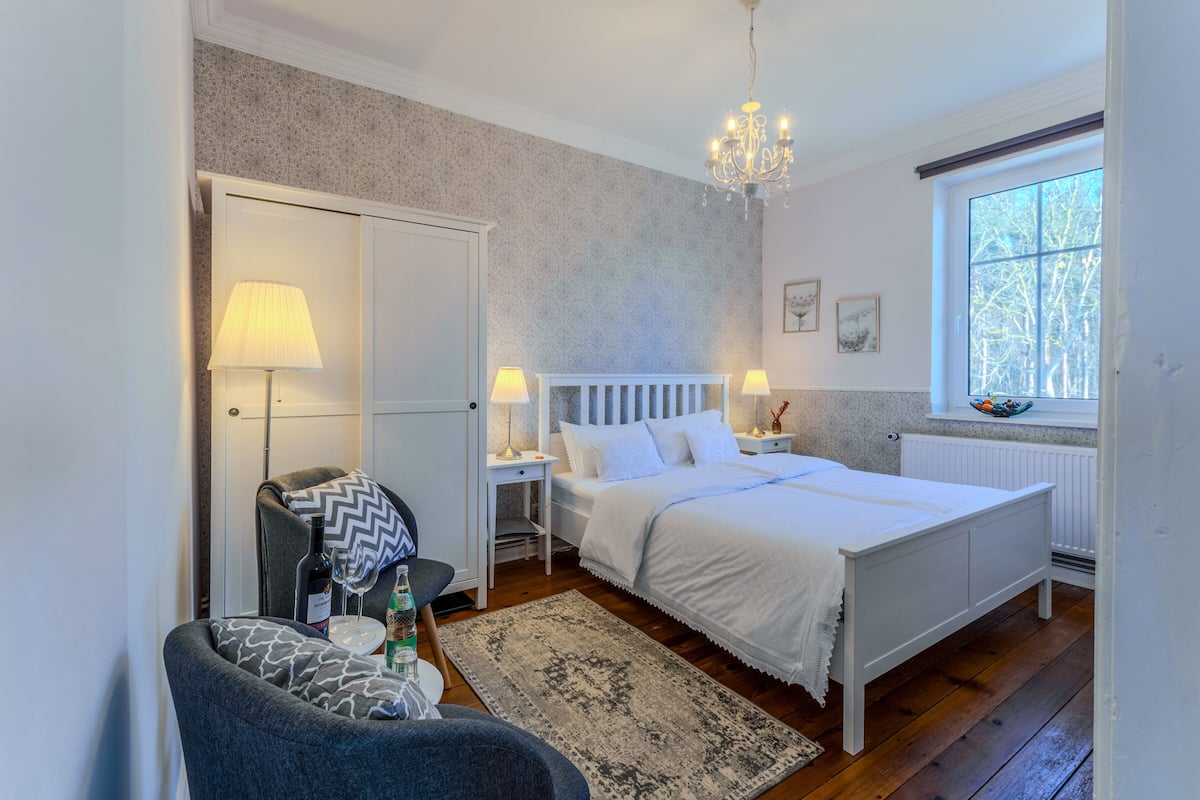 Un dormitorio con cama, mesitas de noche, una silla, candelabro, ventana y radiador.