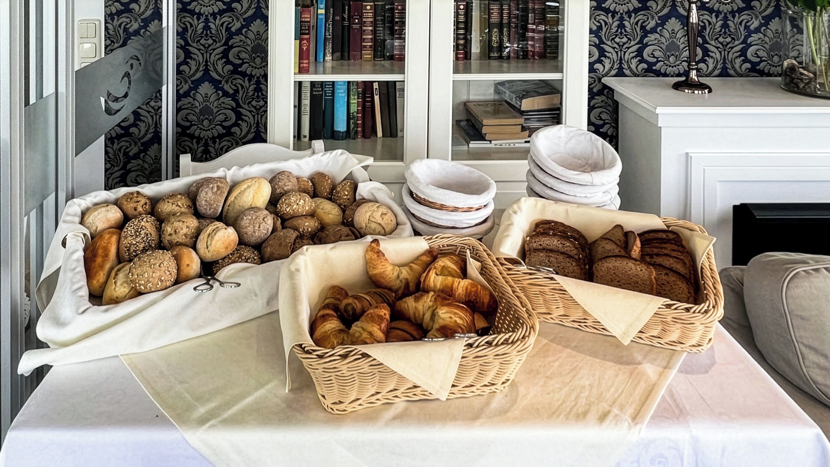 Ein Frühstücksbuffet mit verschiedenen Brotsorten und Gebäck, ein Bücherregal mit Büchern und eine dekorative Vase.