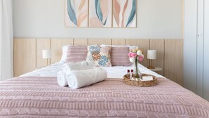Cama bem arrumada com colcha rosa, almofadas brancas e uma almofada decorativa com padrão azul e laranja. Há duas luminárias de cabeceira, um vaso com flores e um cesto com artigos de higiene pessoal na cama.