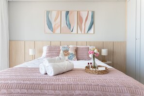 Cama bem arrumada com colcha rosa, almofadas brancas e uma almofada decorativa com padrão azul e laranja. Há duas luminárias de cabeceira, um vaso com flores e um cesto com artigos de higiene pessoal na cama.