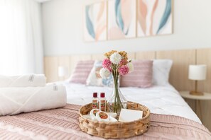 Een netjes opgemaakt bed met een roze deken, een rieten dienblad met toiletartikelen en een bloemenvaas, en een nachtkastje met een lamp.