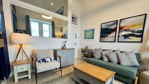 A modern living room with a sofa, a coffee table, a side table, and a lamp. There are framed artworks on the wall and a window with curtains. - Warm, Upgraded Top-floor Flat in Finnieston (Glasgow)