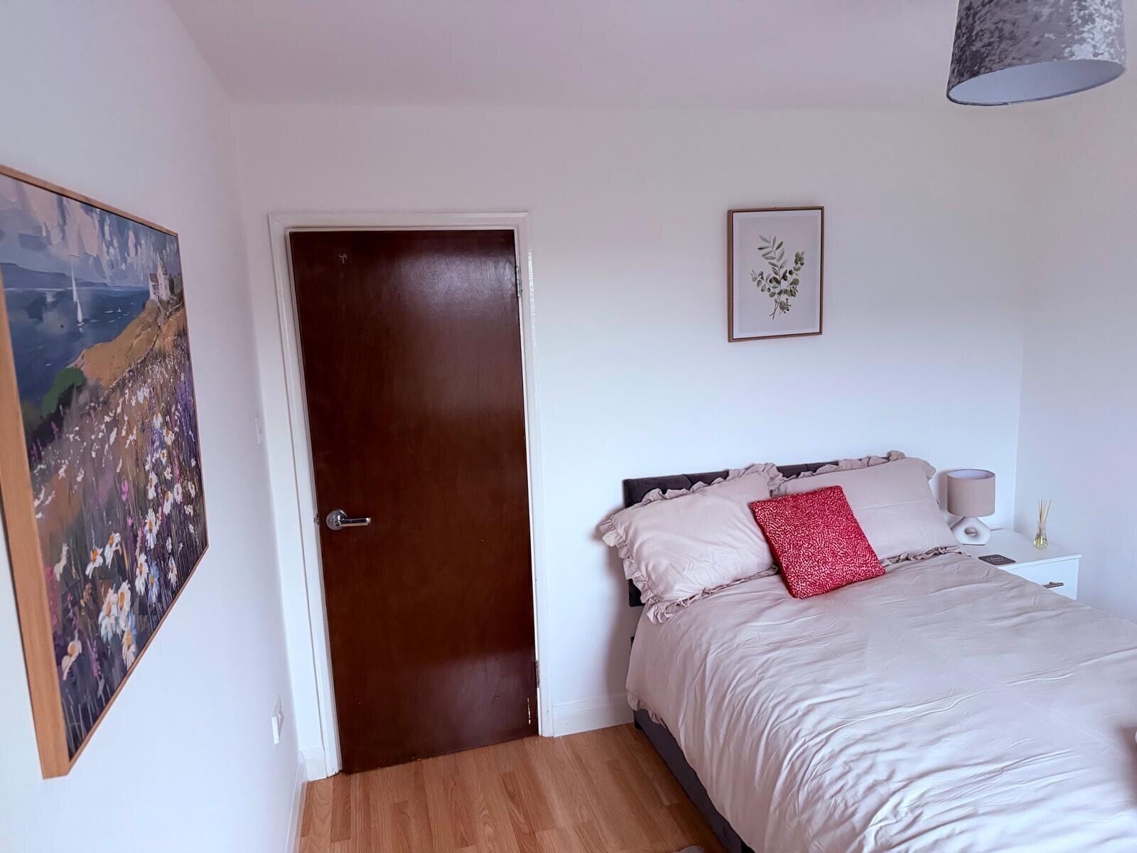 Une chambre avec une porte en bois, un lit avec un oreiller beige et rouge, une table de chevet avec une lampe, et un tableau encadré au mur.