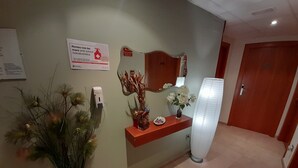 A reception area with a sign indicating first aid services, a wall-mounted first aid kit, a potted plant, a decorative vase, and a tall standing lamp. - HOTEL AVENIDA (El Morell)