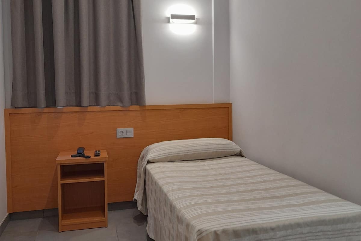 A small, neatly arranged bedroom with a single bed, a bedside table with a lamp, and a window with curtains.
