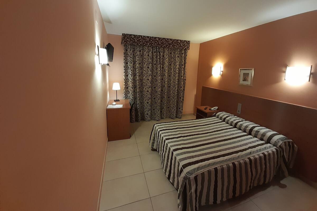 A hotel room with two beds, a desk, a chair, a lamp, a telephone, and patterned curtains.