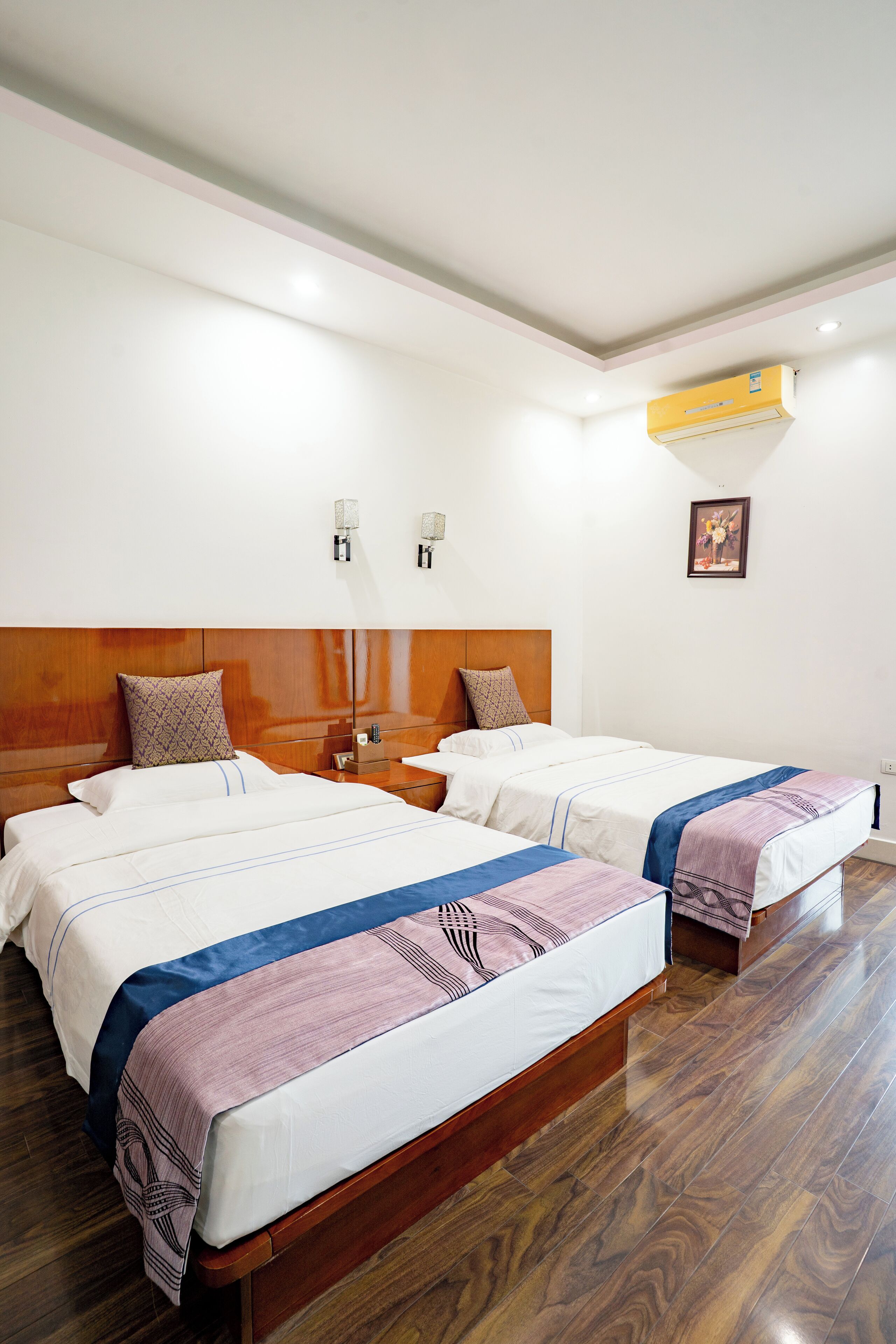 Een hotelkamer met twee bedden, houten hoofdborden, een nachtkastje en een airconditioner aan de muur, met een ingelijste foto.