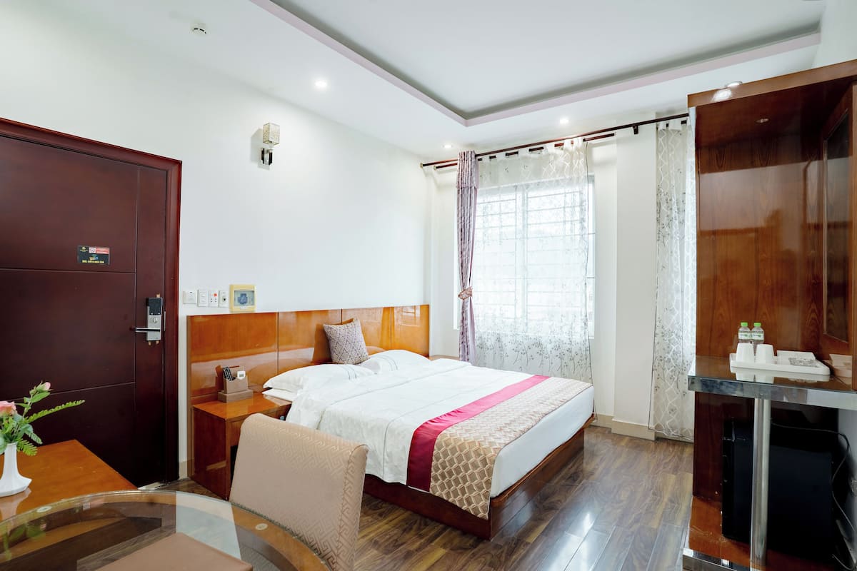 A hotel room with a wooden bed, a desk with a chair, a small table with a vase, and a wardrobe.