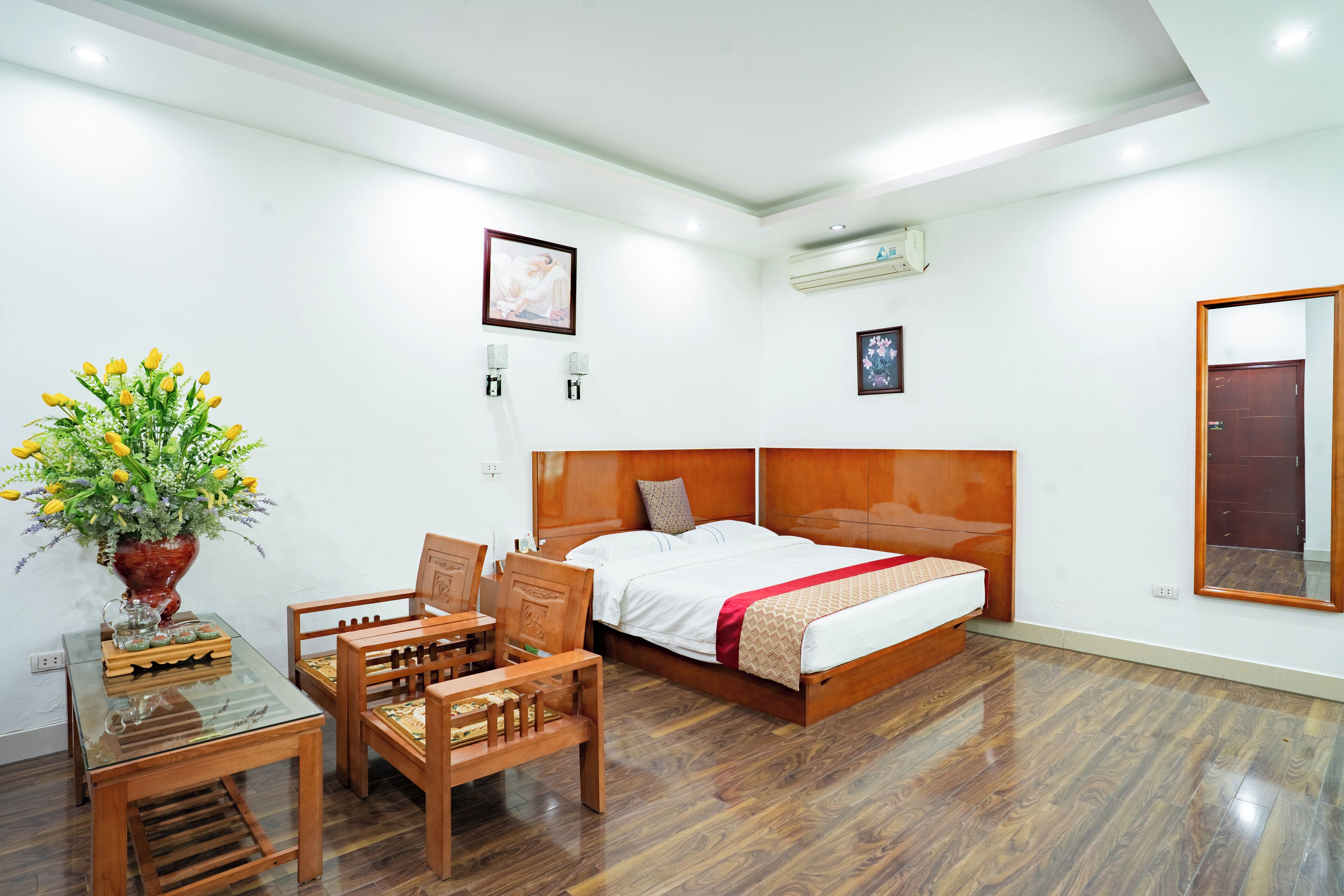 A hotel room with a wooden bed, a desk with a chair, a potted plant, and a mirror on the wall.