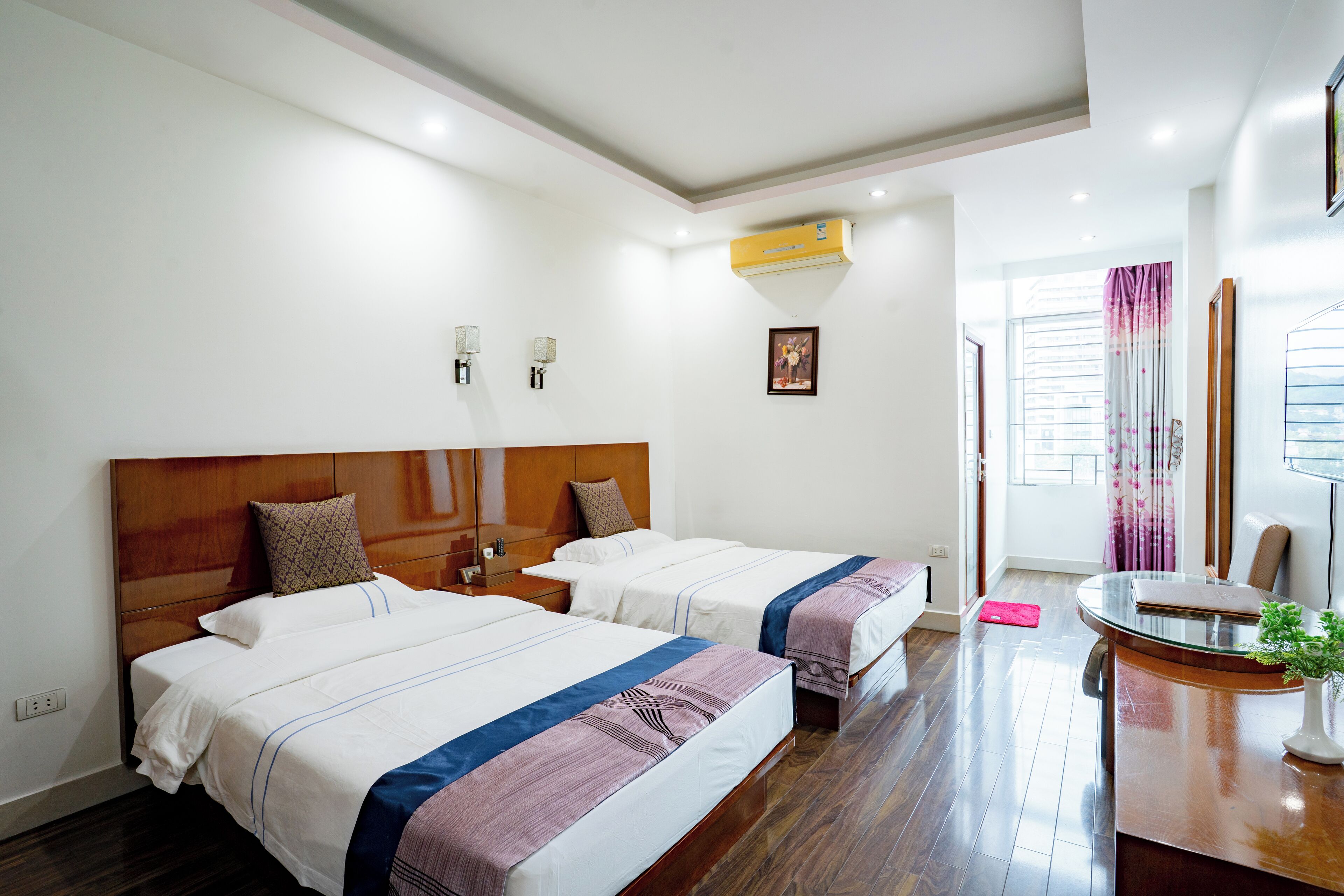 Habitación de hotel con dos camas, cabecera de madera, un escritorio con un florero y una ventana con cortinas.