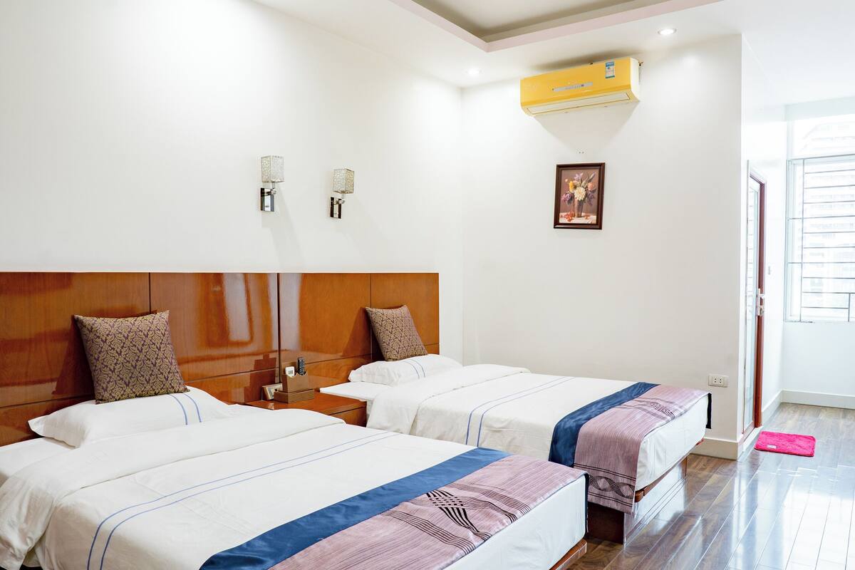 A hotel room with two beds, wooden flooring, a ceiling light, a wall-mounted air conditioner, and a door with a window.