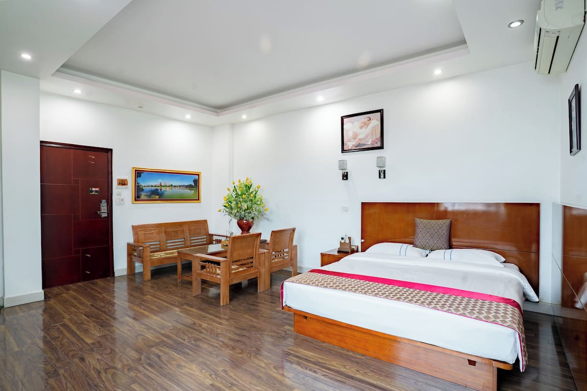 A hotel room with a wooden bed, a wooden desk, a chair, a small table, and a potted plant.
