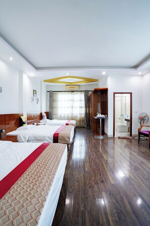 A hotel room with three beds, a wooden floor, a bathroom with a shower, and a fan.