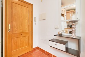 Una puerta de madera, una pared blanca con un espejo, un mueble pequeño y un piso de baldosas.