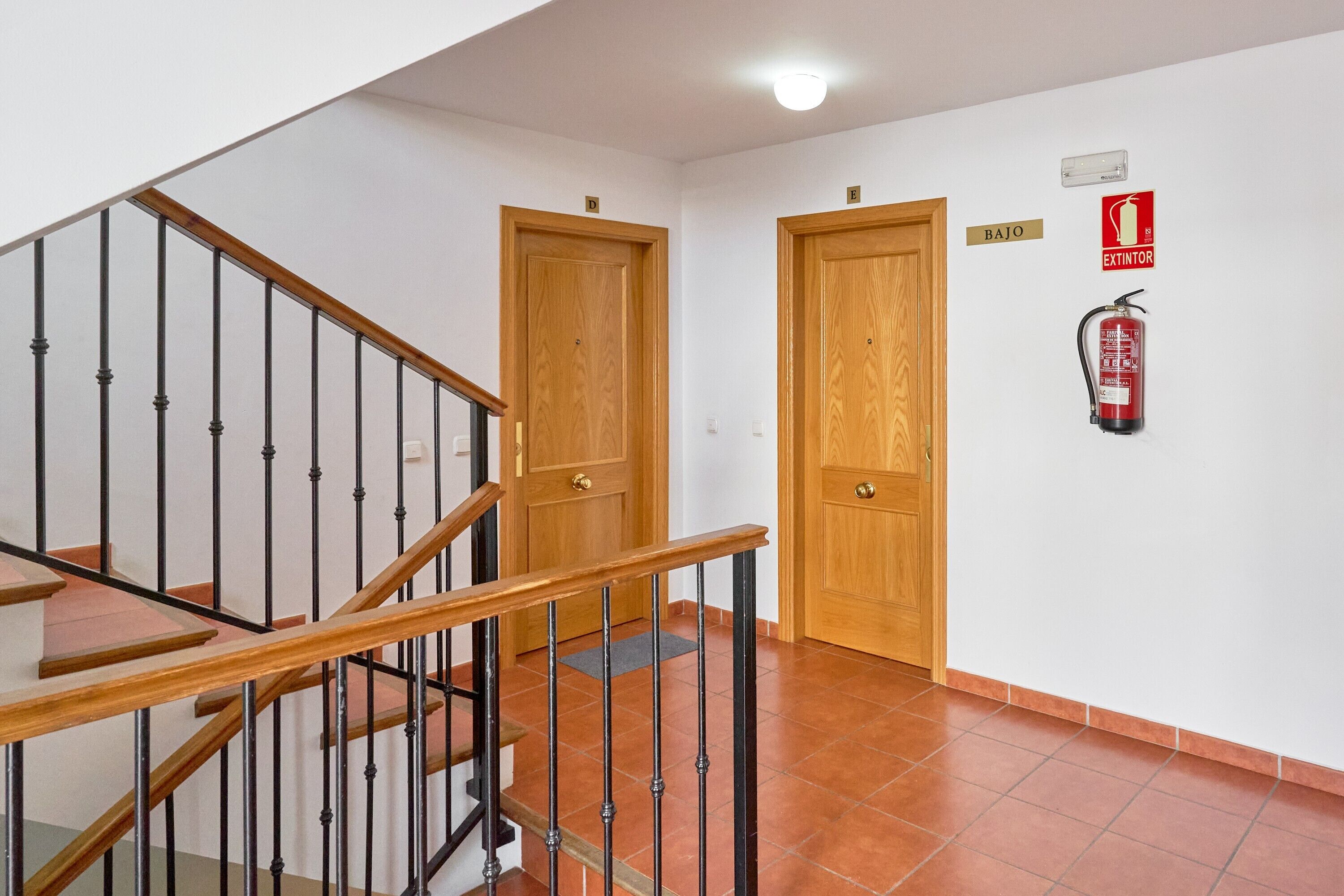 Een trap met houten leuningen en zwarte balustrades, twee houten deuren, een brandblusser aan de muur en een lamp aan het plafond.