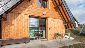 A wooden cabin with a large sliding glass door, a dining area with a table and chairs, and a small outdoor seating area. - Le Crozet, quality cottage with panoramic mountain views (Plateau-des-Petites-Roches)