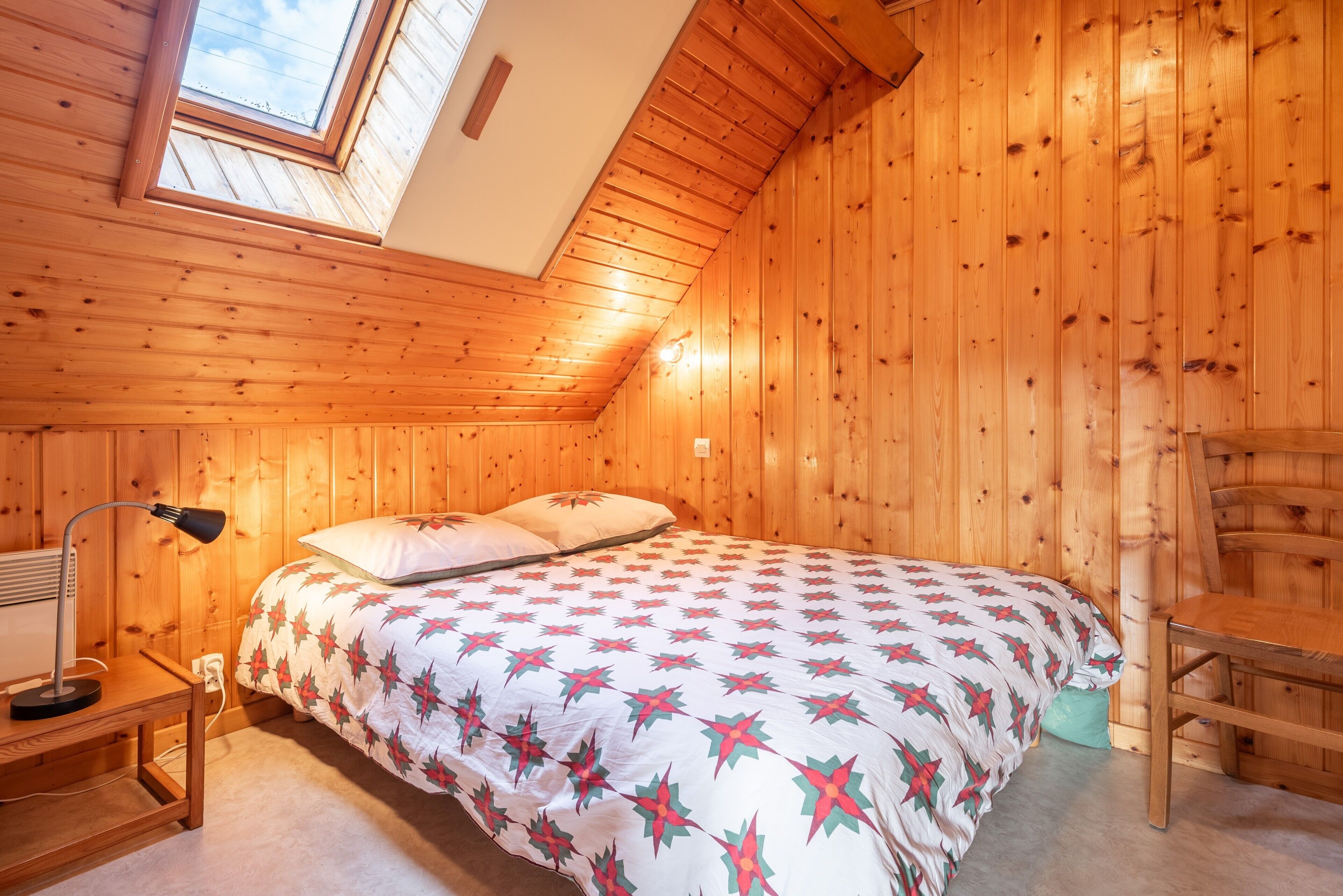 A wooden-paneled room with a bed, a desk, a chair, and a skylight.