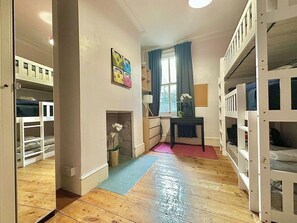 Ein Zimmer mit Etagenbetten, einem kleinen Tisch mit Lampe, einem Fenster mit Vorhängen und einem Wandbild.