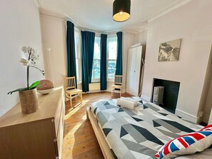 A bedroom with a bed, a chair, a small table with an orchid, a fireplace, and a window with curtains. - Large Property in the Heart of London (London)