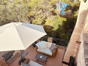 Un balcon avec un parasol blanc, des meubles en osier et une vue sur des arbres et une étendue d’eau bleue.