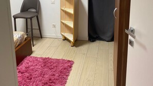 A small, sparsely furnished room with a wooden floor, a grey chair, a small wooden cabinet, and a pink rug. - Peaceful 2-bedroom apartment in charming Balerna perfect for relaxing stays (Balerna)