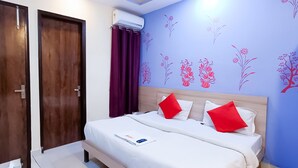 A hotel room with a wooden door, a bed with white bedding and red pillows, and a wall with floral wall art. - Roomshala Hotel Rose Residency (New Delhi)