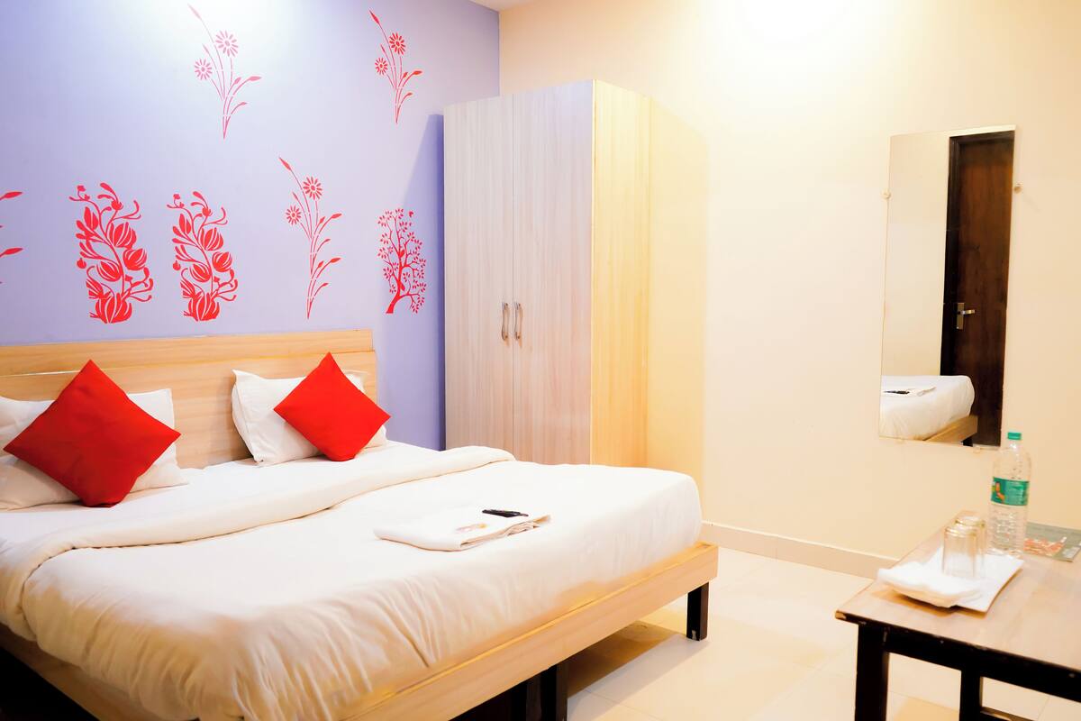 A neatly made bed with red pillows, a wardrobe, a mirror, and a small table with a bottle and glasses.