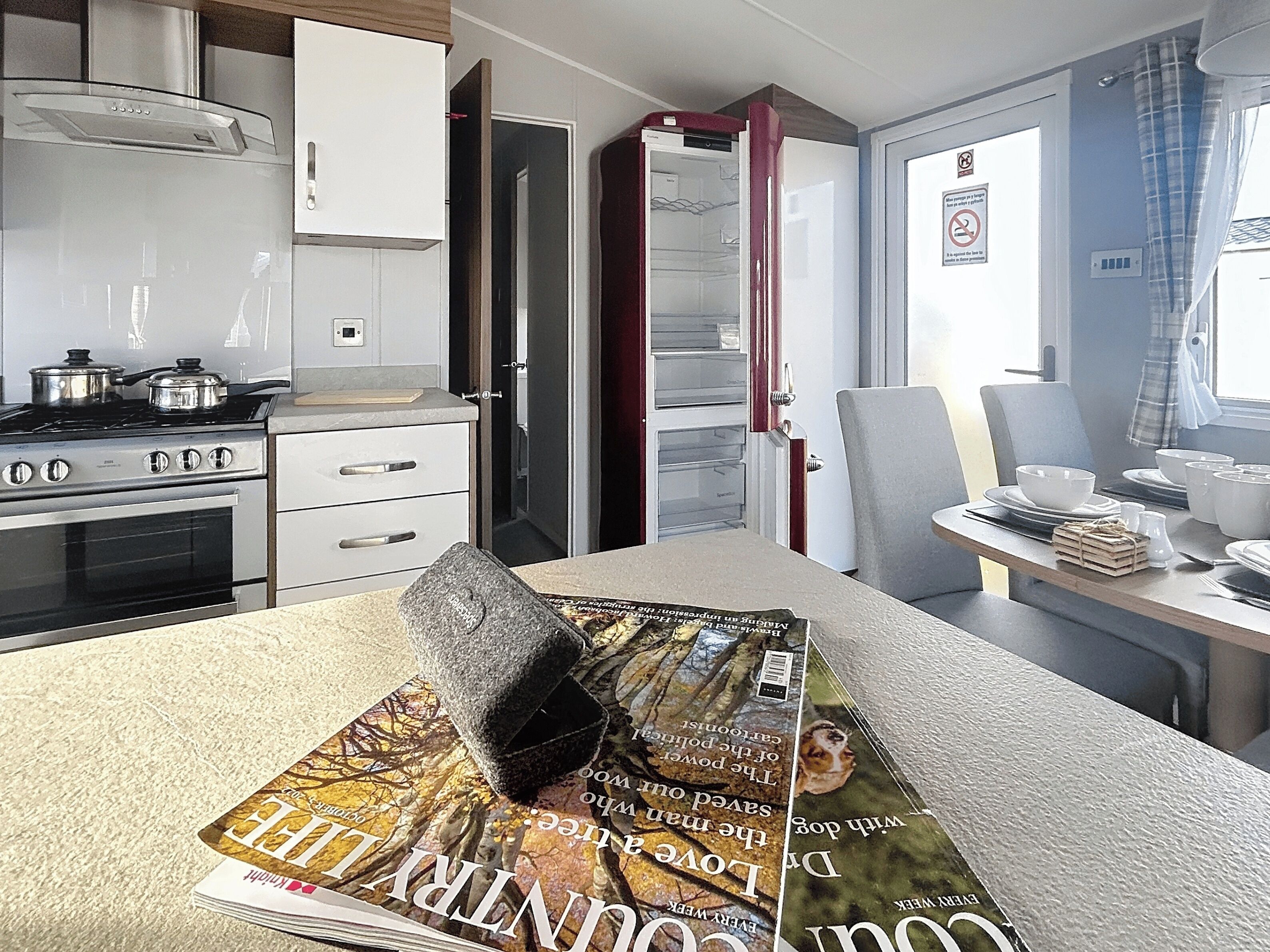 A compact kitchen with a stove, oven, and white cabinets. A dining table with a magazine and a cushion is in the foreground.