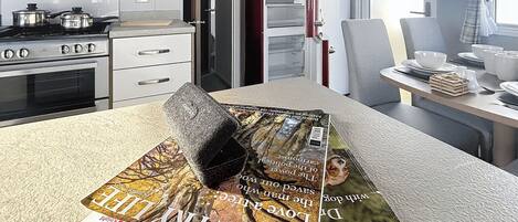 A compact kitchen with a stove, oven, and white cabinets. A dining table with a magazine and a cushion is in the foreground.