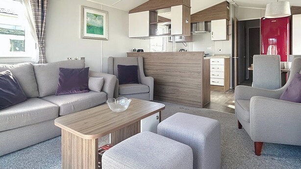 A modern living room with a sofa, armchairs, a coffee table, and a kitchen area with cabinets and a red appliance. - Premium Lodge - Sleeps 8 - Seton Sands Beach (Prestonpans)