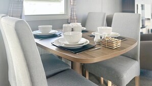 A dining area with a wooden table set for four, grey upholstered chairs, white tableware, and a small stack of books on the table. - Premium Lodge - Sleeps 8 - Seton Sands Beach (Prestonpans)