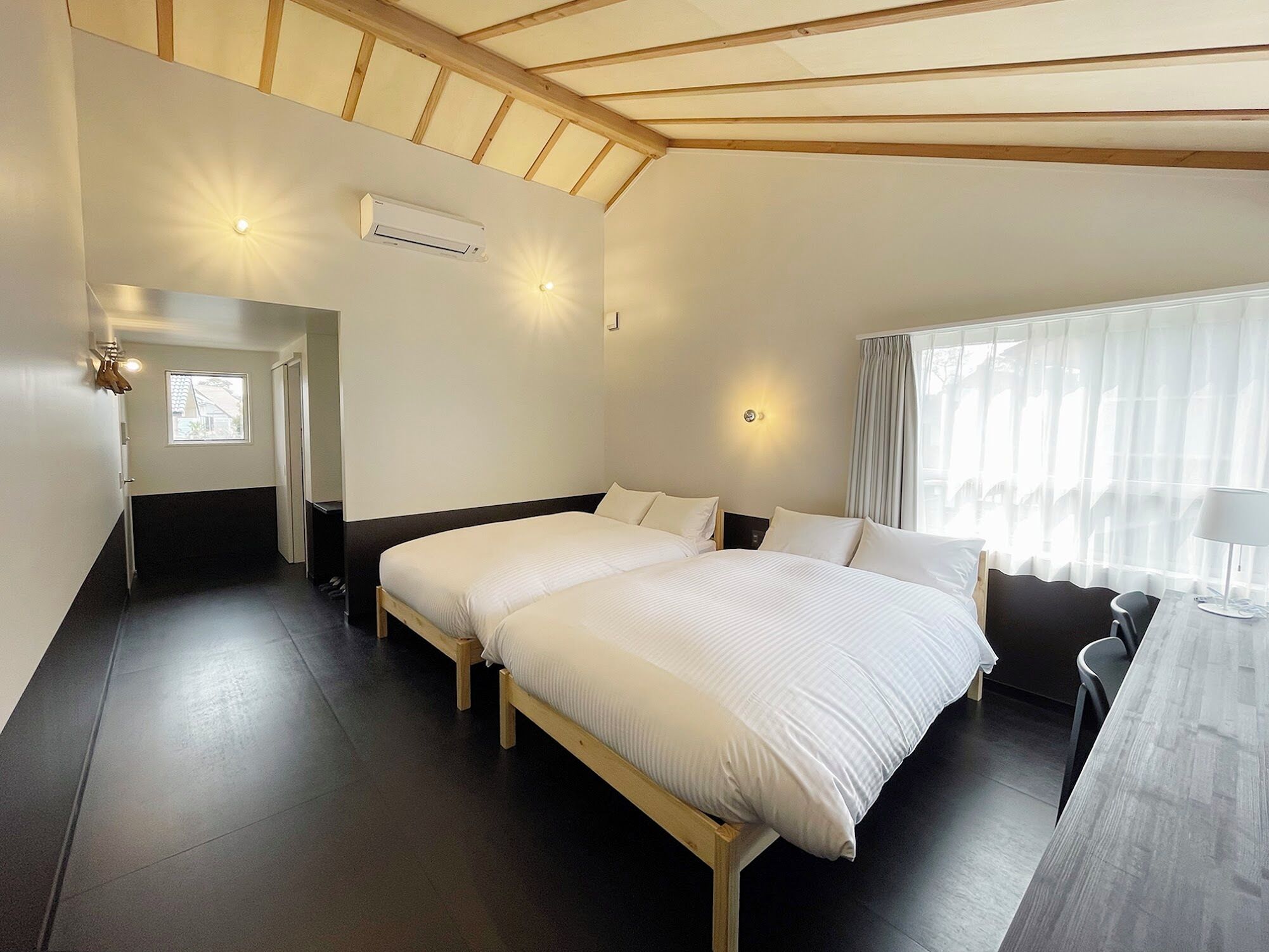 A hotel room with a large bed, a desk with a chair, a window with curtains, and an air conditioning unit.