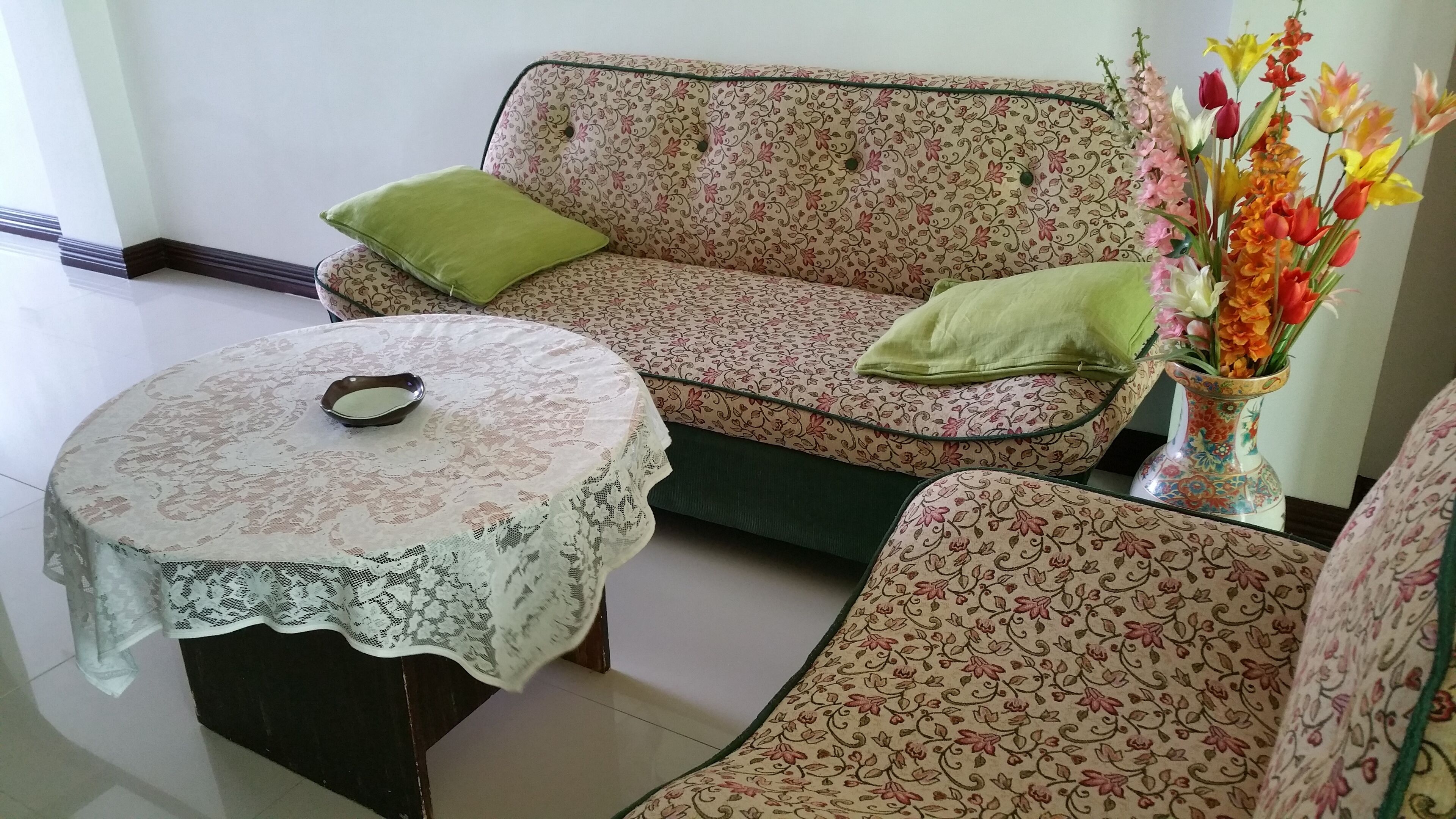 Een woonkamer met een bloemenbank, een bijpassende fauteuil, een ronde tafel met een kantkleed en een vaas met bloemen.