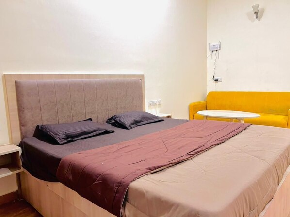 A hotel room with a bed, a yellow sofa, a small table, and a wall-mounted light fixture. - Stay by Sunstone (Greater Noida)