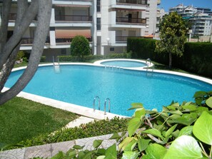A swimming pool surrounded by greenery, with a building in the background. - Family apartment with pool - 6 guests (Gandía)