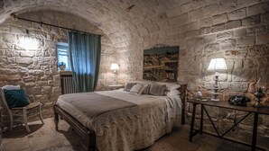 Un dormitorio con pared de piedra, una cama con cubrecama de encaje, una silla, una mesa con lámpara y un cuadro en la pared.