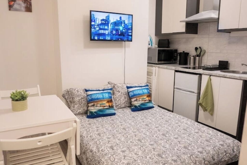A small, well-lit room with a bed, a TV mounted on the wall, a kitchenette, and a dining area.