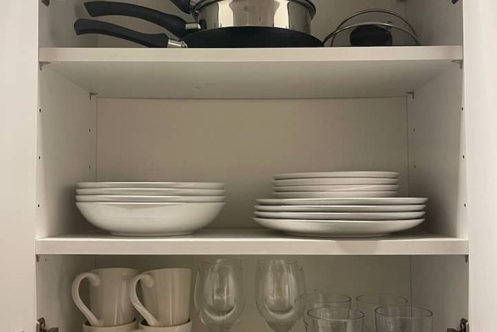 Una estantería con platos y tazones blancos, y una repisa inferior con tazas y copas de vino.