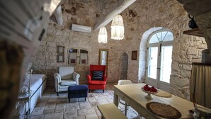 Une pièce rustique aux murs de pierre, avec un plafond haut, une table en bois sur laquelle se trouve un panier de fruits, un fauteuil rouge, un fauteuil blanc et un banc.