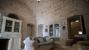 Una habitación rústica con pared de piedra, un armario blanco con puertas de cristal, un sofá con manta y una silla.