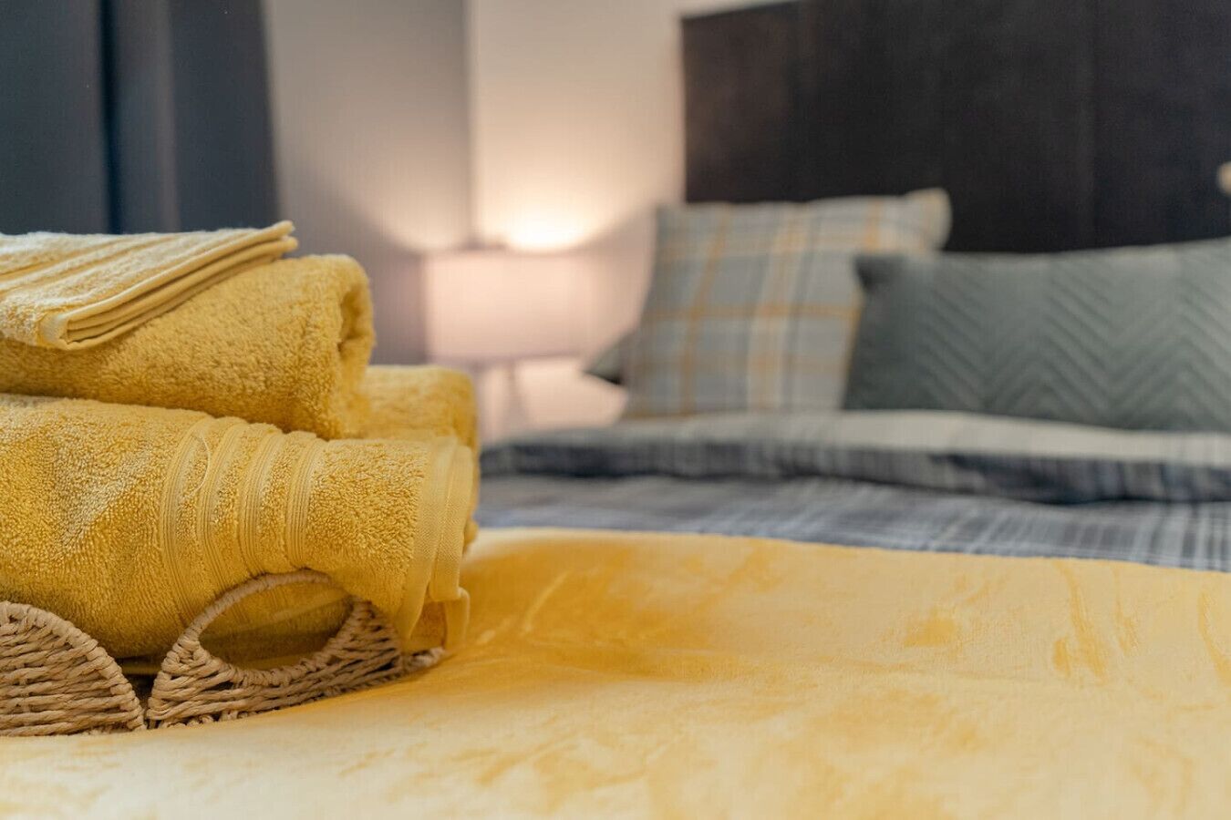 Cama bem arrumada com colcha amarela, toalha amarela dobrada e cesta de vime sobre a cama.