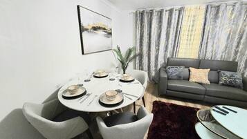 A dining area with a round table set for four, surrounded by grey chairs. A grey sofa with cushions is against the wall, and a large window with curtains is visible.