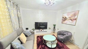 A modern living room with a grey sofa, a round coffee table, a grey armchair, and a flat-screen TV on a white stand. - Gem Place - A Home Away From Home (Hornchurch)