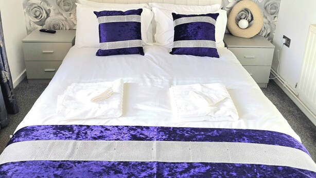 A neatly made bed with white and purple linens, a headboard with a floral pattern, and a bedside table with a lamp. - Gem Place - A Home Away From Home (Hornchurch)