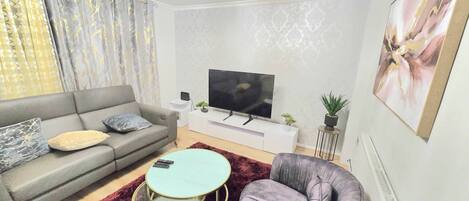 A modern living room with a grey sofa, a round coffee table, a grey armchair, a TV on a white stand, and a large abstract painting on the wall.