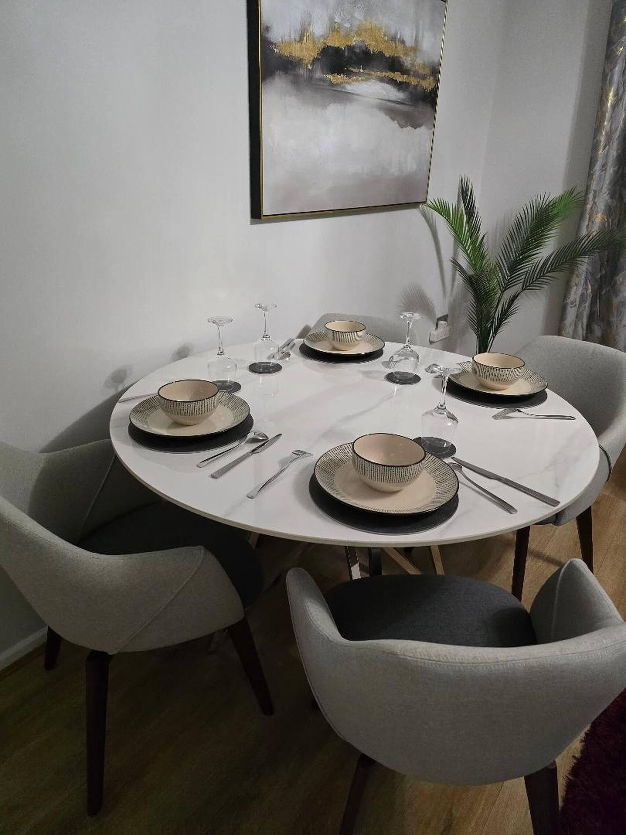 A dining table set for four with a white tablecloth, black and beige plates, and silverware. There are four chairs around the table, and a potted plant is visible in the background.