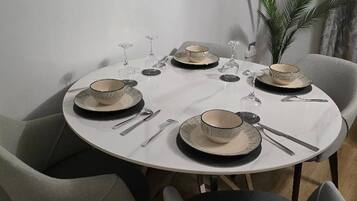 A dining table set for four with a white tablecloth, black and beige plates, and silverware. There are four chairs around the table, and a potted plant is visible in the background.