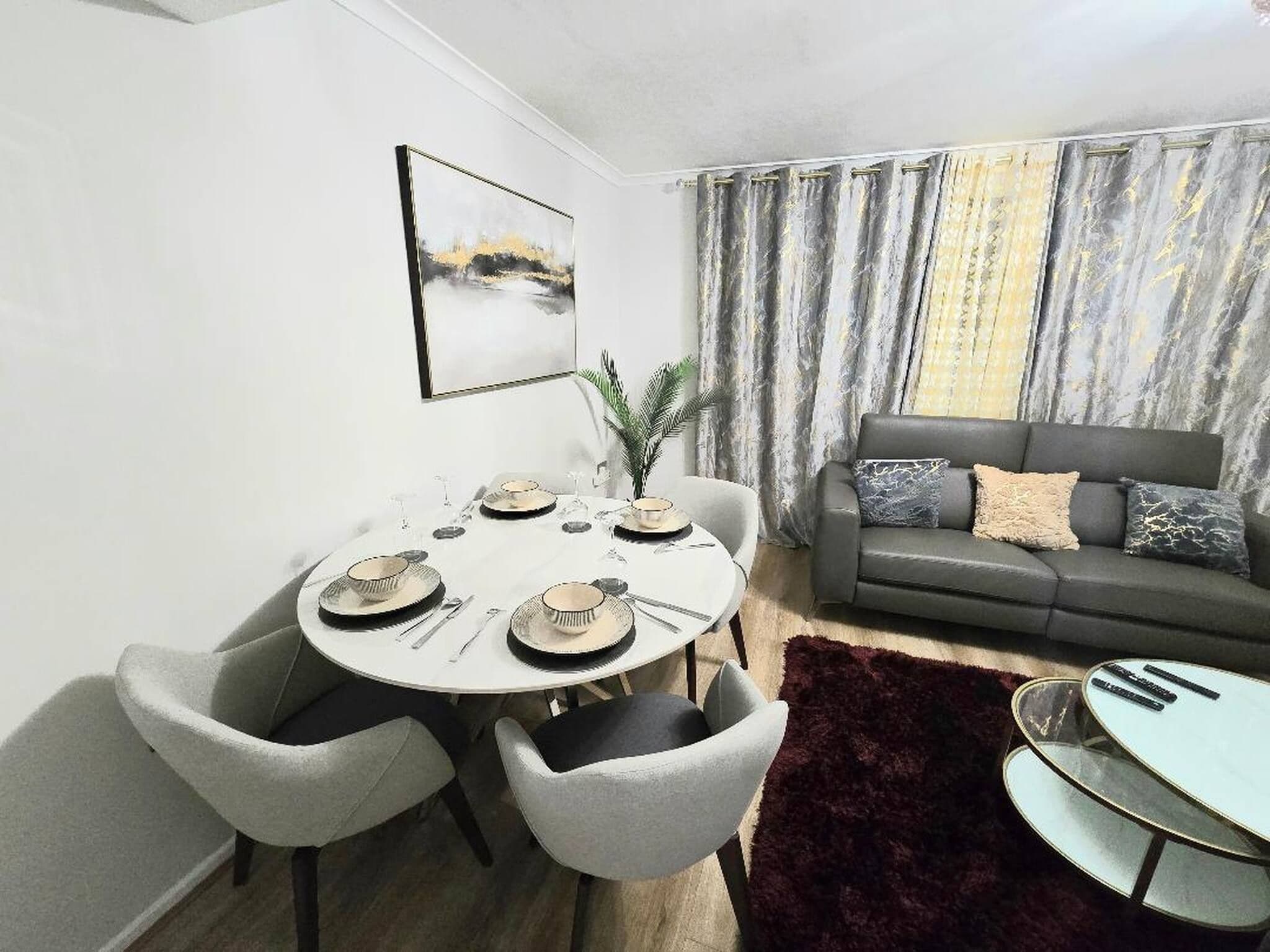 Un coin repas avec une table ronde dressée pour quatre personnes, entourée de chaises grises. Un canapé gris avec des coussins est adossé au mur, et une grande fenêtre avec des rideaux est visible.