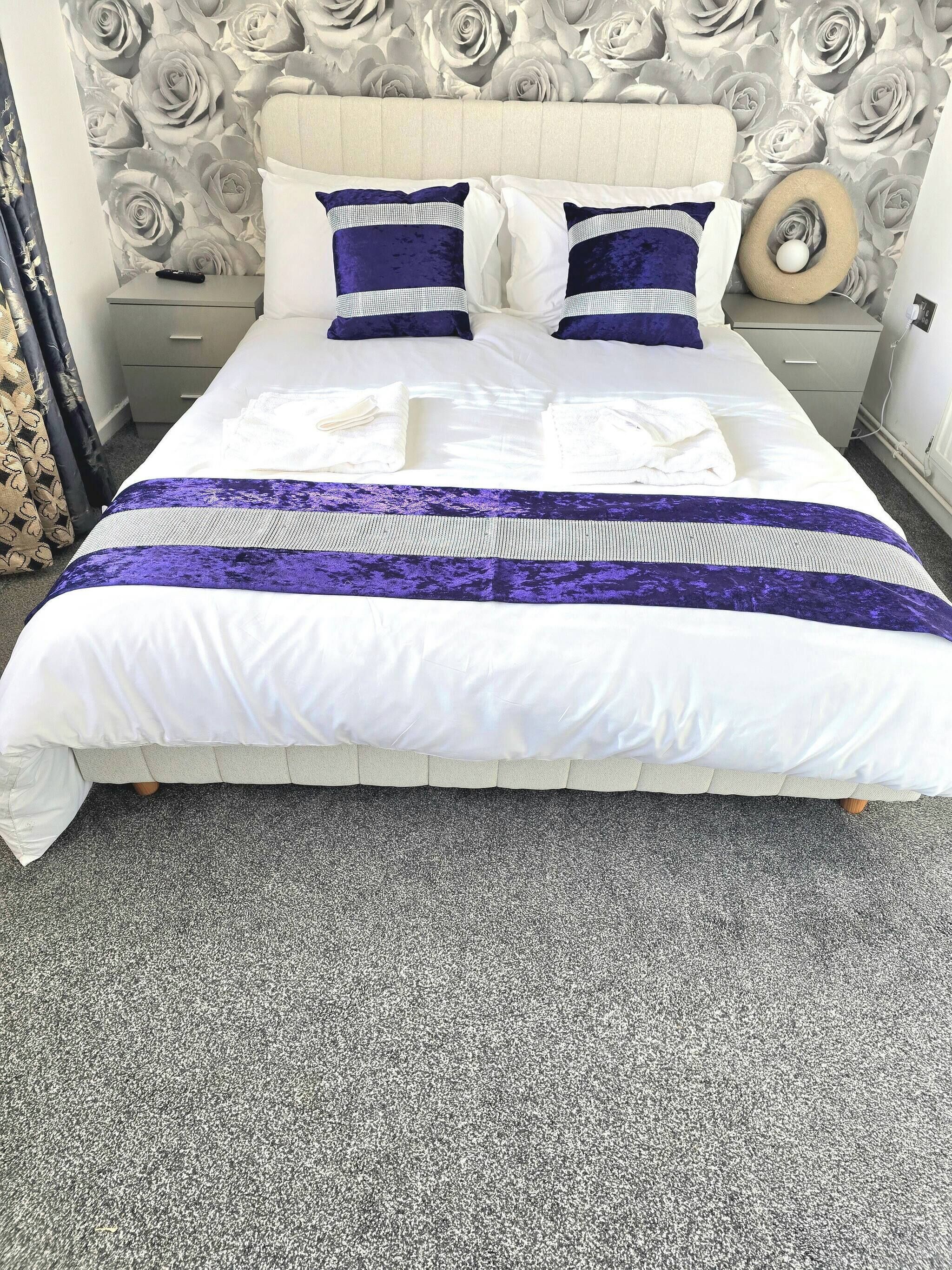 Un lit bien fait, recouvert d’une couvre-lit blanche et violette, avec une tête de lit ornée d’un motif floral et un tapis gris.
